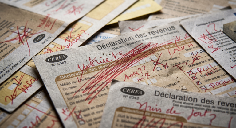 Gros plan sur une pile désordonnée de formulaires fiscaux français CERFA 'Déclaration des revenus', couverts de ratures agressives, de gribouillages et de points d'interrogation au stylo rouge. L'éclairage est direct et cru.