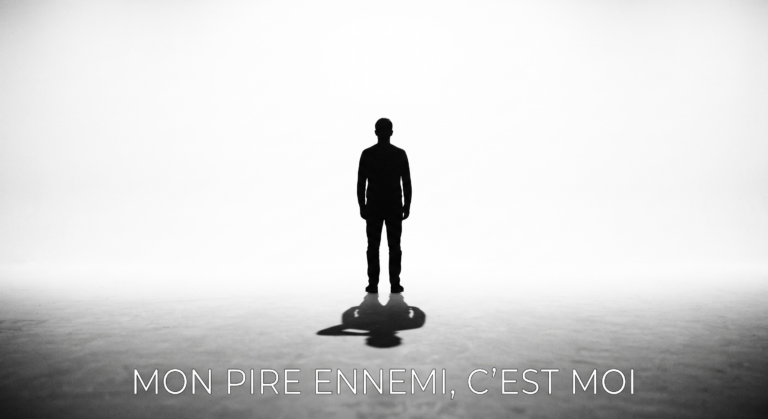 Une photographie conceptuelle en noir et blanc montrant la silhouette d'une personne de dos, seule, debout face à une lumière vive. Son ombre est projetée vers le sol. Le texte français "MON PIRE ENNEMI, C'EST MOI" est affiché au bas. Cette image symbolise la lutte intérieure liée aux erreurs d'investisseur dues à la psychologie.